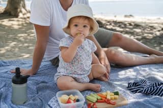 Niño pequeño comiendo un picnic sencillo a la sombra en la playa con supervisión de un adulto