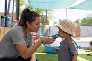 Educadora ofreciendo agua a un niño pequeño con gorro en un patio sombreado de guardería en verano