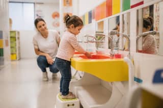 Peque lavándose las manos en un lavabo adaptado de guardería con supervisión de educadora