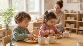 Dos pequeños comiendo en una mesa baja de guardería con supervisión cercana de la educadora en un aula luminosa