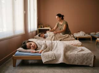 Peque durmiendo en una cuna del aula de escuela infantil mientras una educadora ajusta la mantita con suavidad