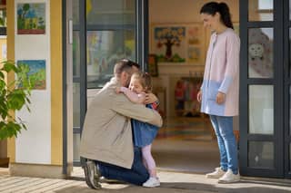 Padre despidiéndose con calma de su hija en la puerta de una escuela infantil mientras la educadora recibe al peque el primer día de adaptación