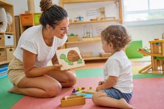 Educadora de ludoteca arrodillada a la altura de un niño pequeño mostrándole un puzzle en una sala de juego luminosa