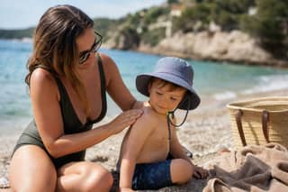 Madre aplicando protector solar a un niño pequeño con gorro en la playa