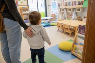Peque explorando un aula de guardería con estanterías bajas, libros y percheros con nombres mientras su progenitor le acompaña