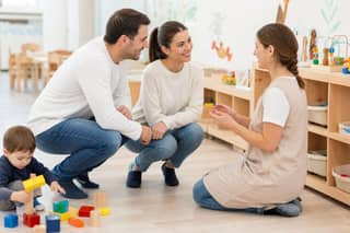 Progenitores hablando con una educadora de guardería a la altura del niño en una sala con juguetes y mobiliario infantil
